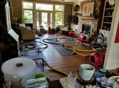 Air movers, hoses and drying equipment set up around the furniture of a living room with a wood floor