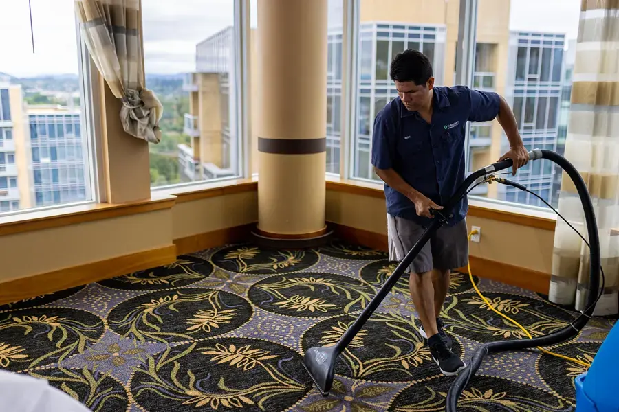 A technician running an extraction hose and wand over a soaked carpet in a bright room